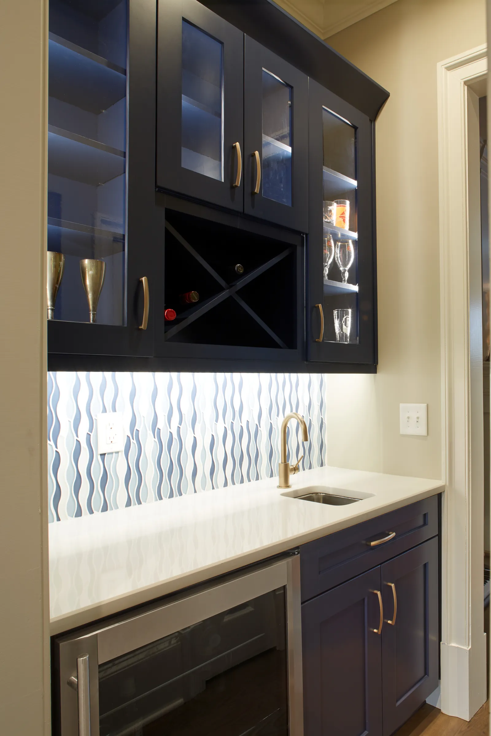 Modern wet bar with navy blue cabinets, wine rack, white countertop, brass faucet, and wave-patterned backsplash.
