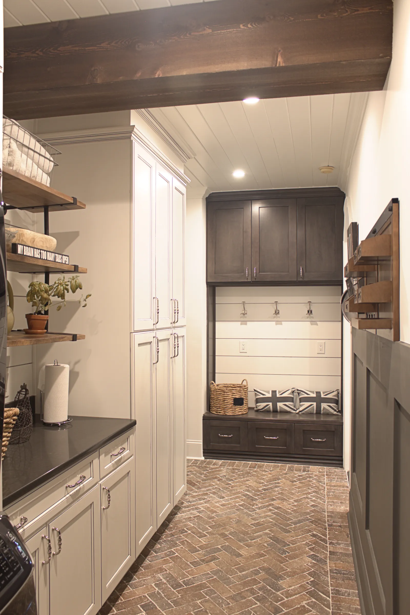 Modern laundry room with dark wood storage bench, white cabinets, herringbone tile floor, and rustic ceiling beam