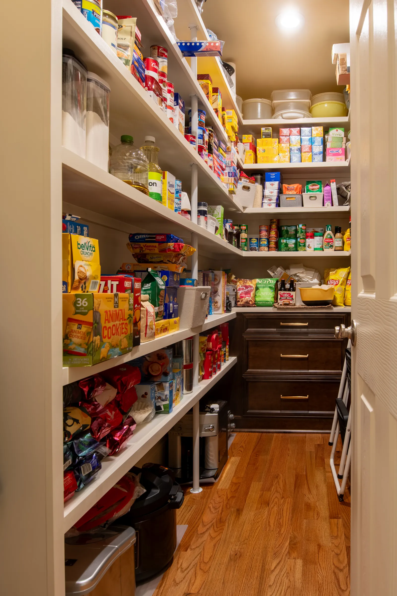 Organized pantry shelves filled with snacks, canned goods, and kitchen supplies in a well-lit storage room.