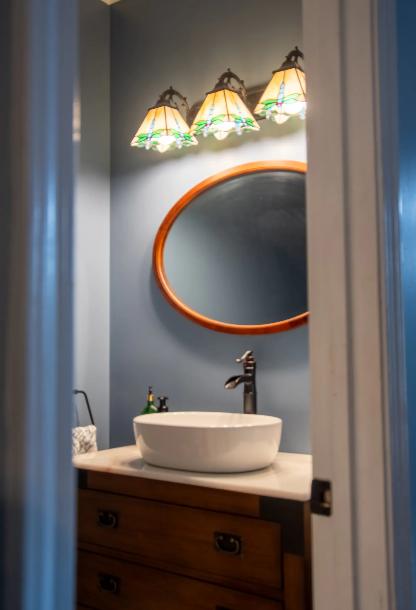 Bathroom vanity with round mirror, vessel sink, bronze faucet, wooden cabinet, and Tiffany-style light fixtures.