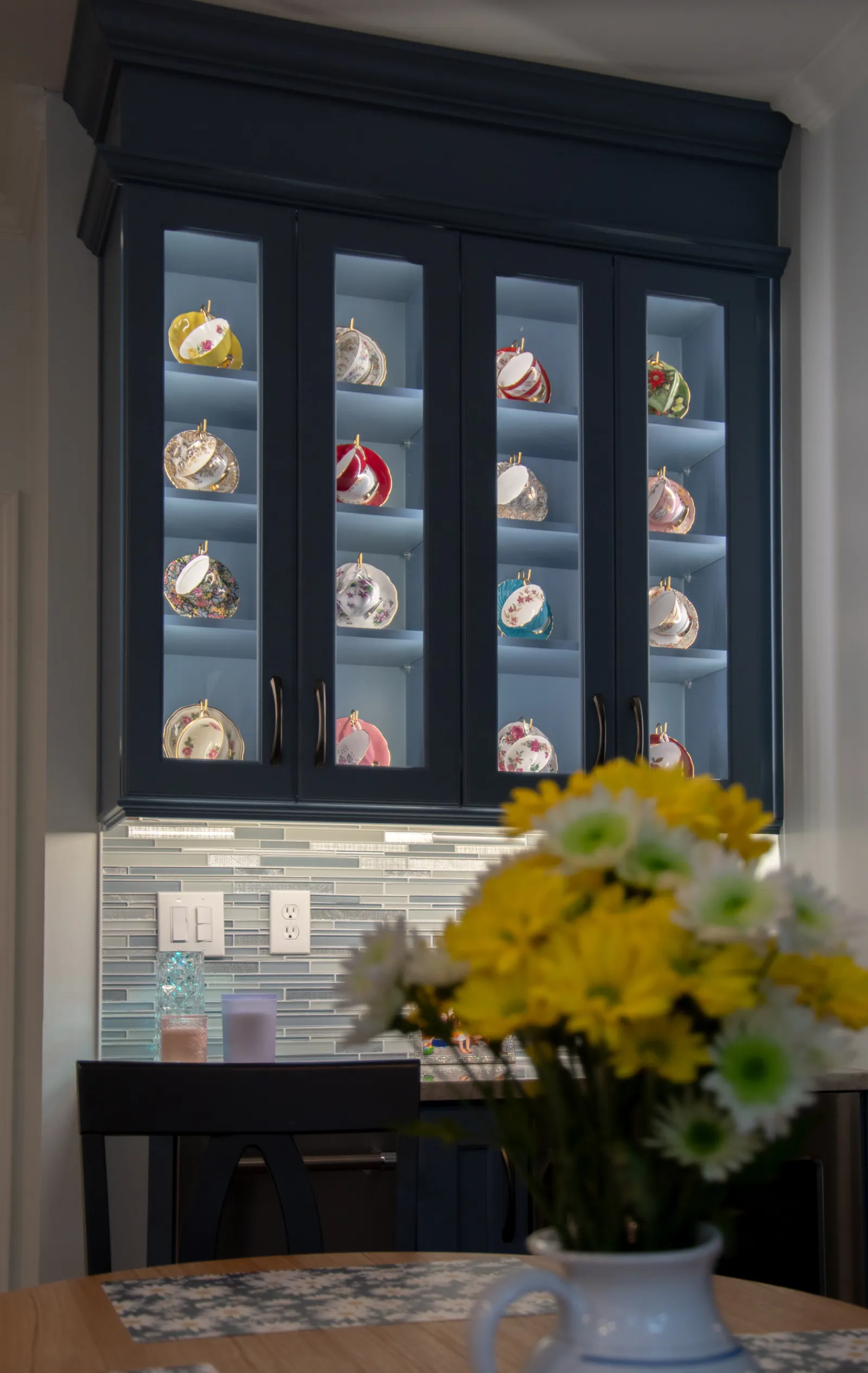 Glass-front cabinet displaying colorful teacups and plates with a vase of yellow and white flowers in foreground