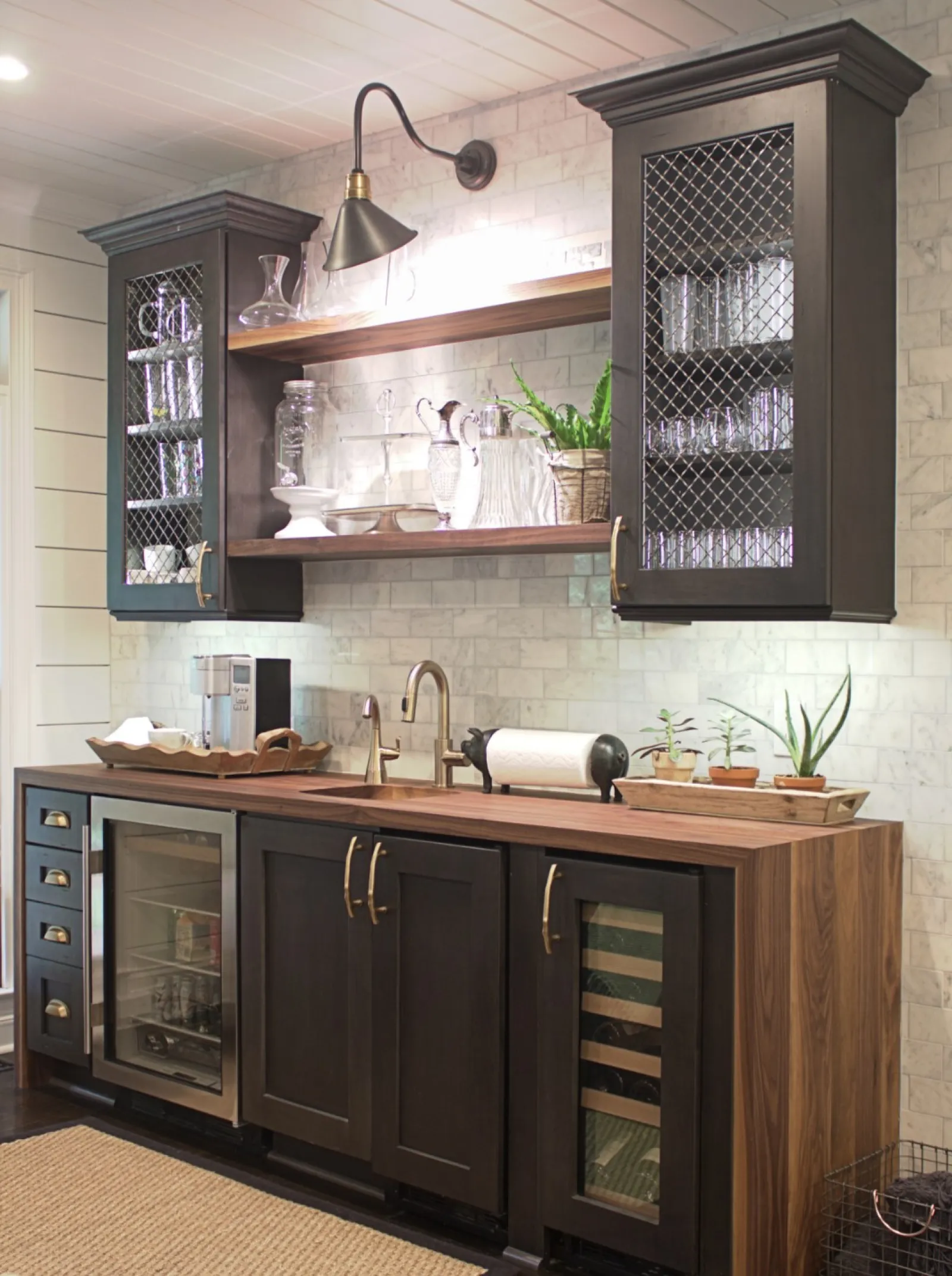 Modern home bar with dark wood cabinets, wine fridge, glassware, and open shelving against white tile wall.