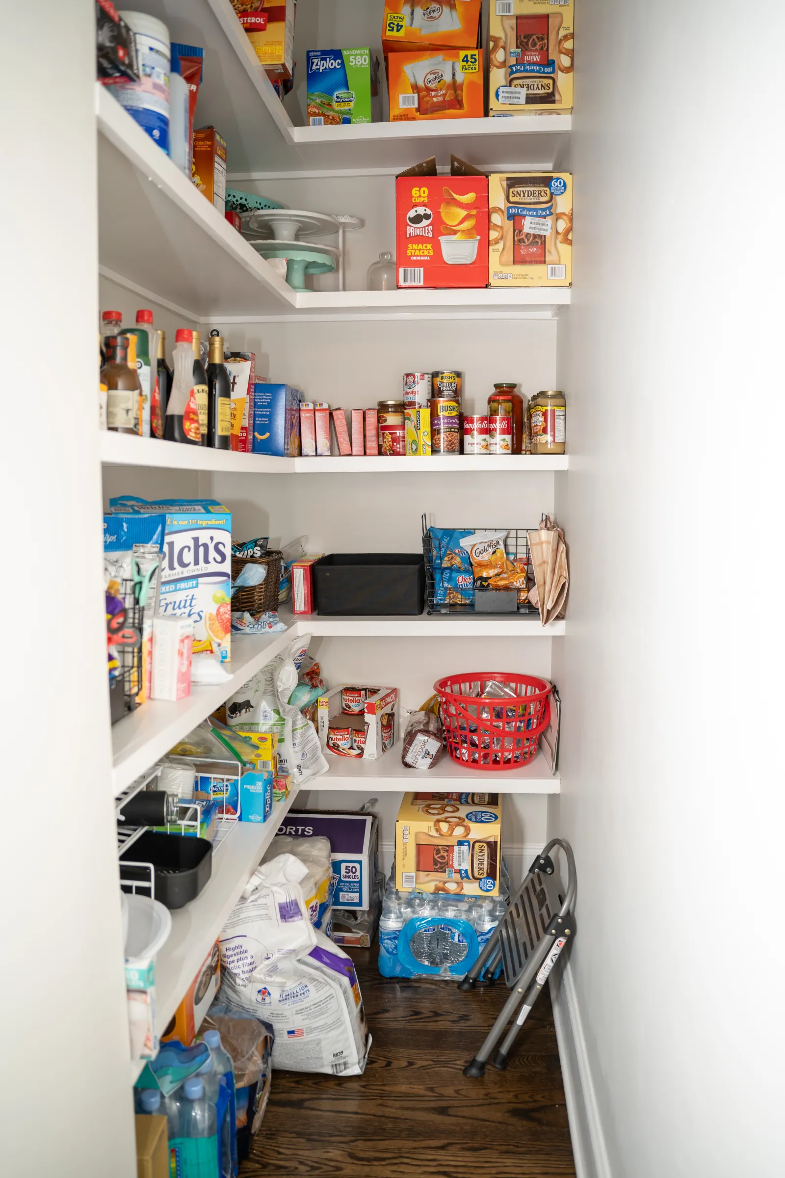 Organized pantry with shelves stocked with snacks, canned goods, bottled drinks, water packs, and kitchen items.