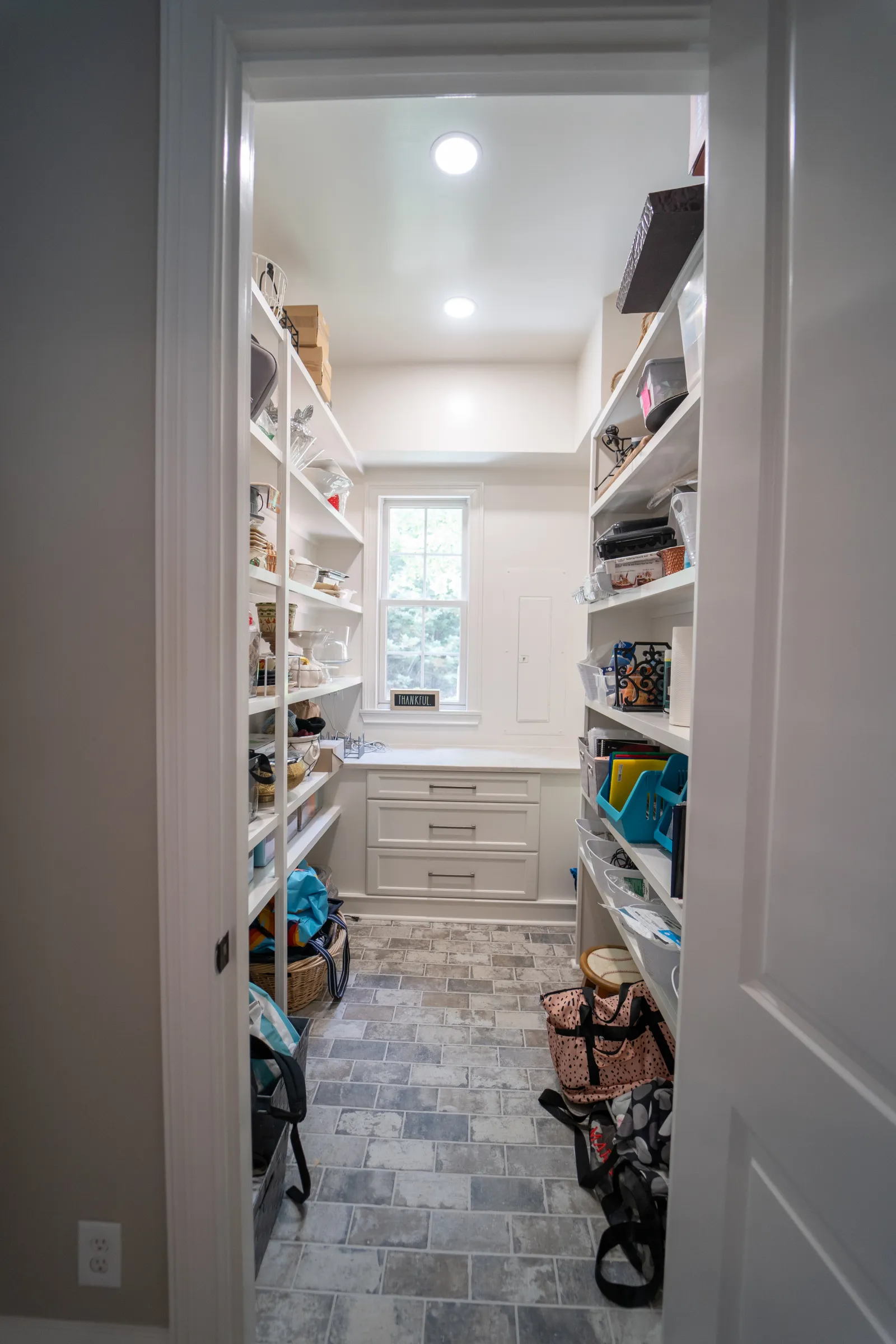 Bright pantry room with white shelves, storage containers, bags, and drawers under a window with natural light.