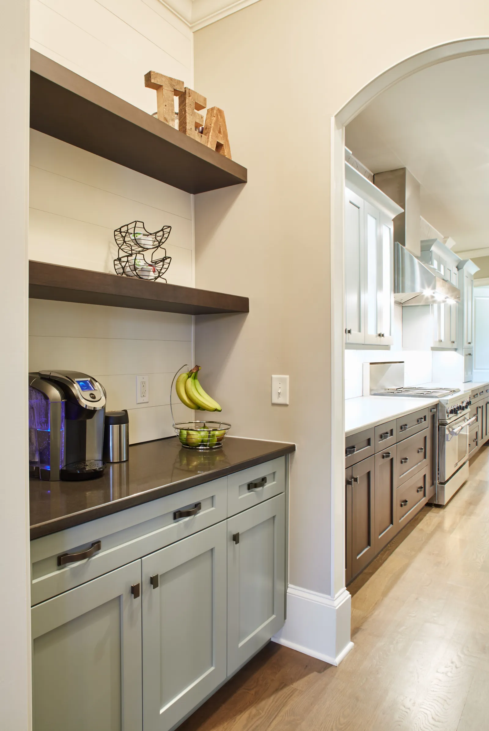 Modern kitchen with gray and wood cabinets, coffee maker, fruit basket, and wooden TEA letters on shelves