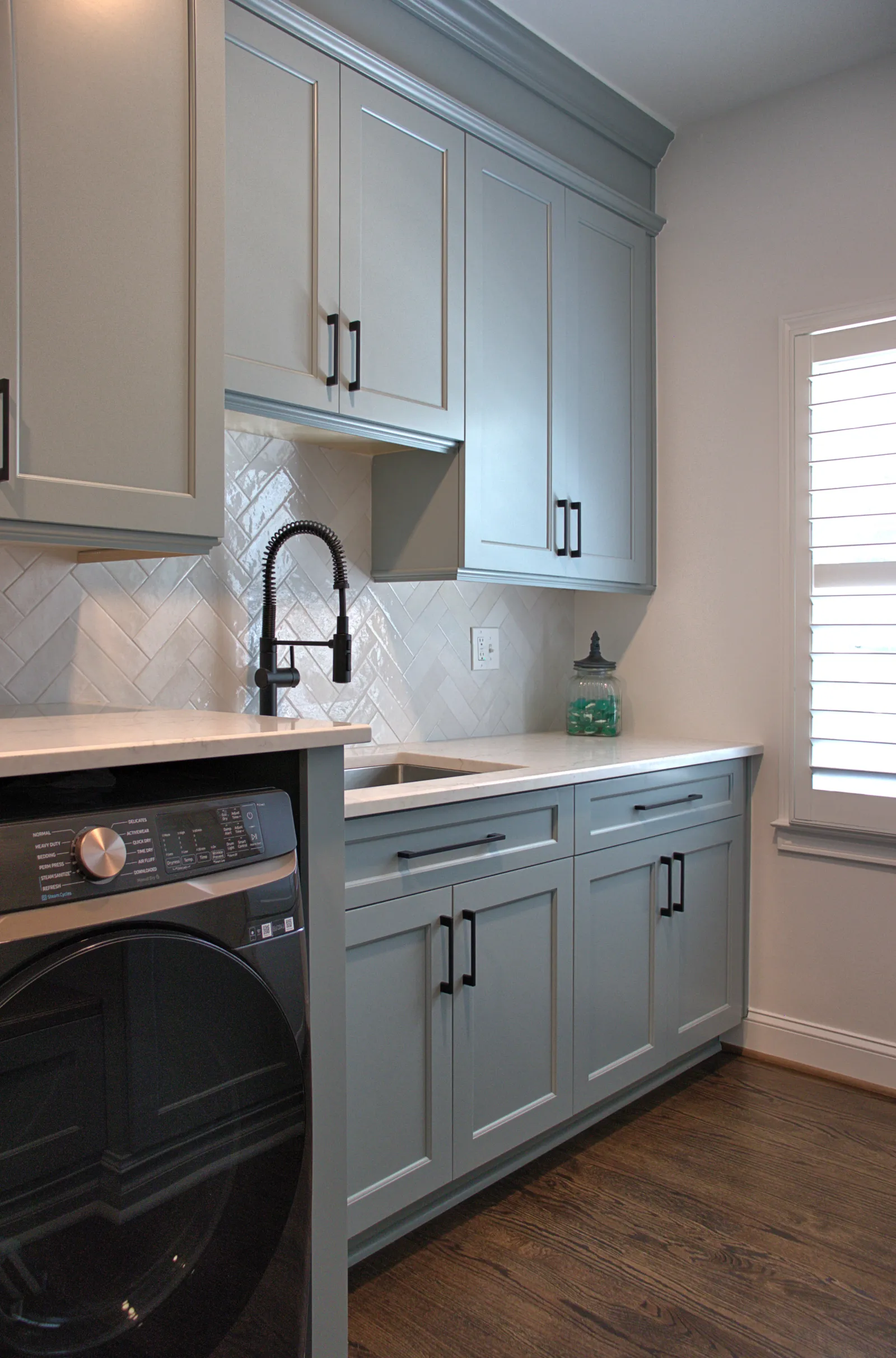 Modern laundry room with gray cabinetry, black hardware, farmhouse faucet, white countertop, and dark wood floor.