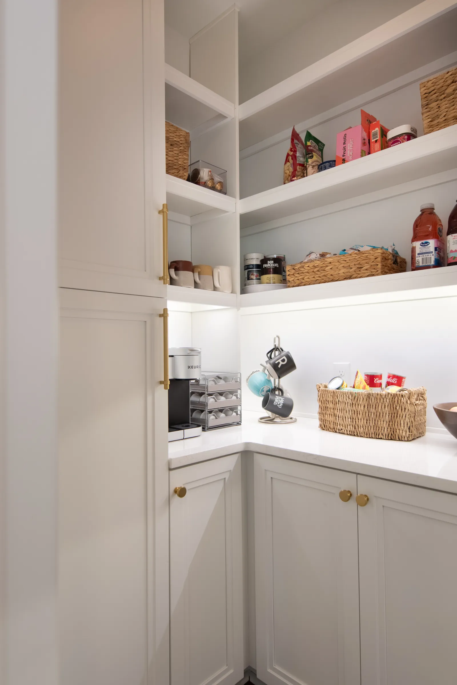 Bright white pantry corner with shelves holding snacks, mugs, coffee machine, and woven baskets.
