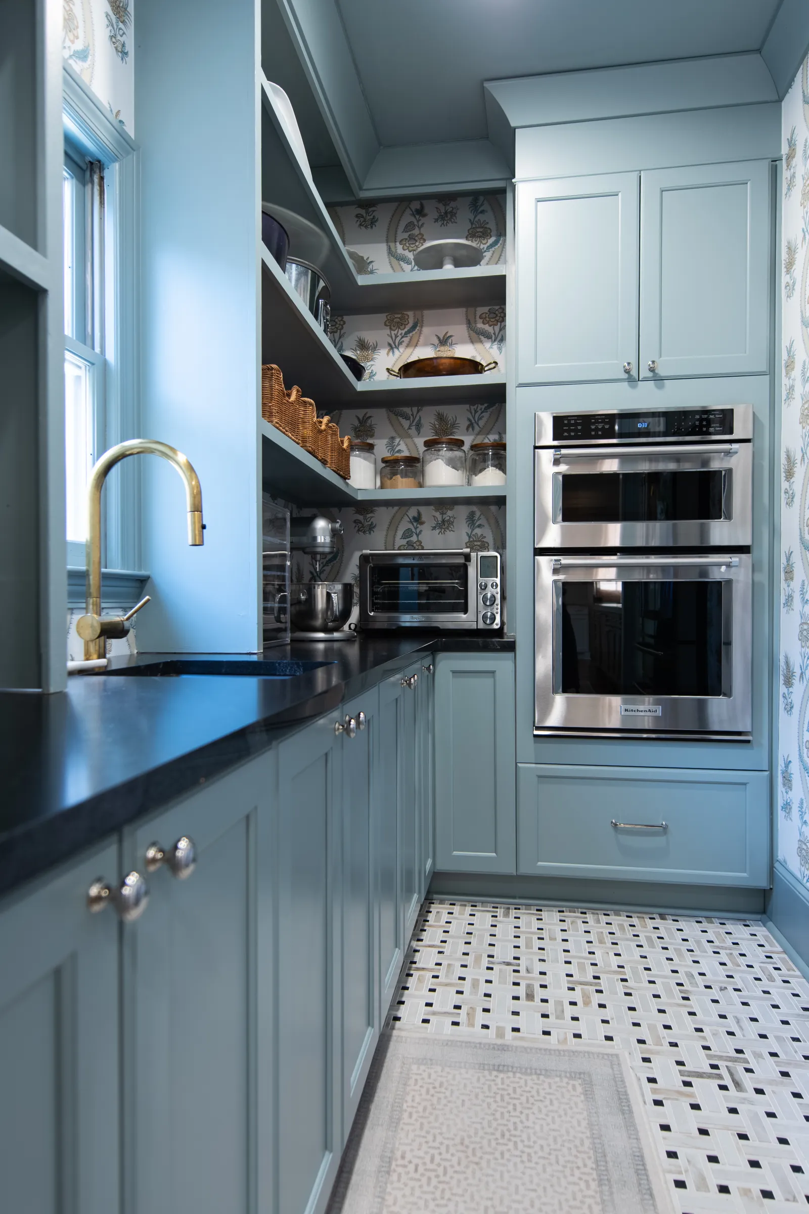 Modern blue kitchen with black countertop, gold faucet, built-in stainless steel double oven, and patterned tile floor.