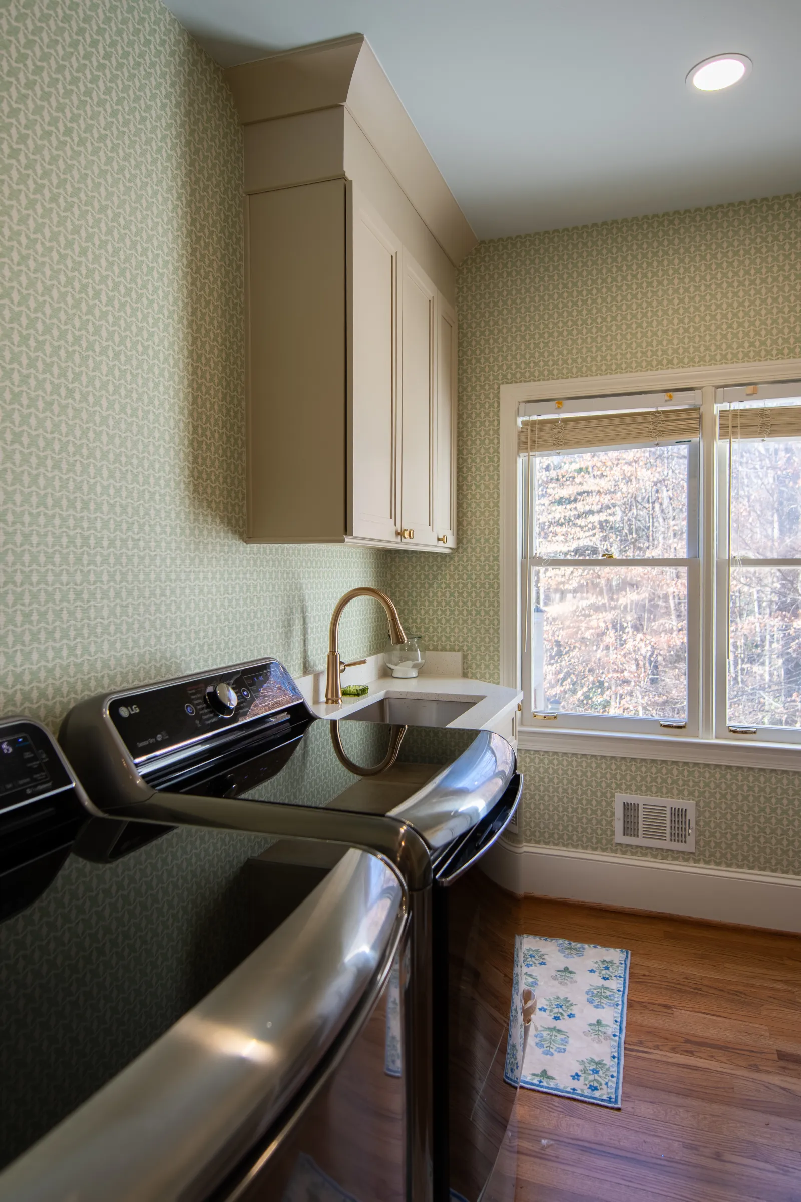 Bright laundry room with modern black washer and dryer, white cabinetry, patterned wallpaper, and large window.