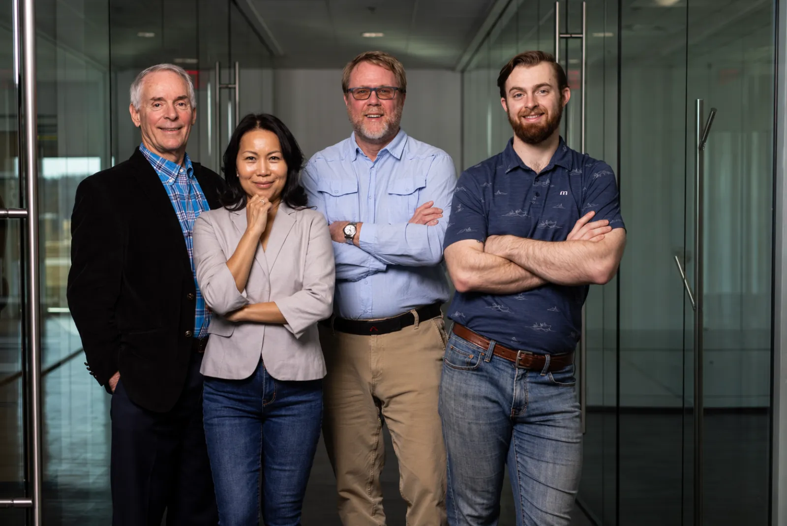 Group of four diverse professionals standing confidently in office with glass walls, smiling and relaxed.