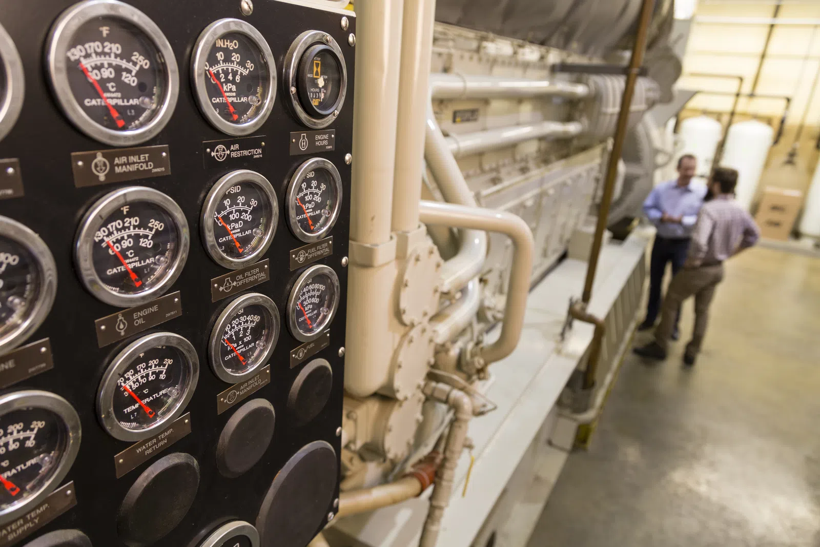 Close-up of industrial control panel gauges with two men discussing by large machinery in a factory setting