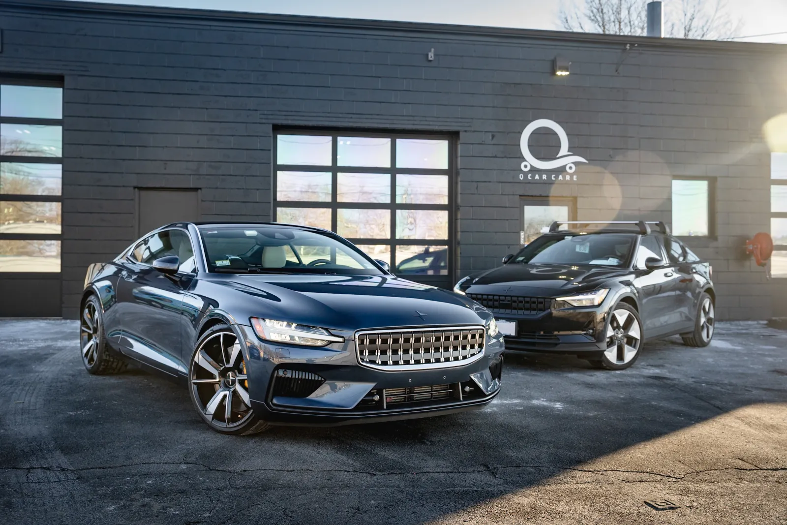 Two sleek dark electric cars parked outside a modern garage with large glass windows and sunlight reflections.