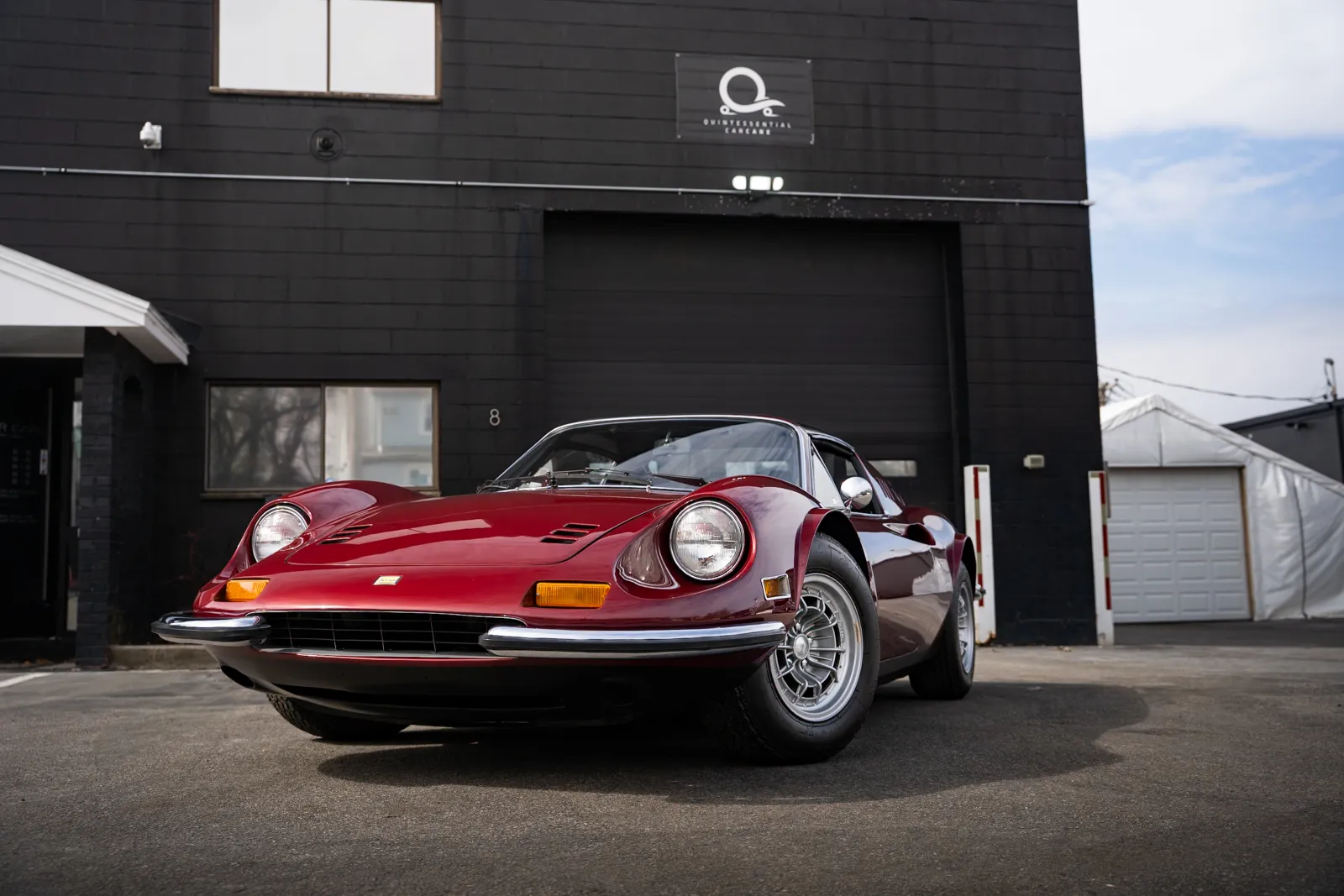Classic red Ferrari Dino parked outside a modern black building on a sunny day with clear skies.