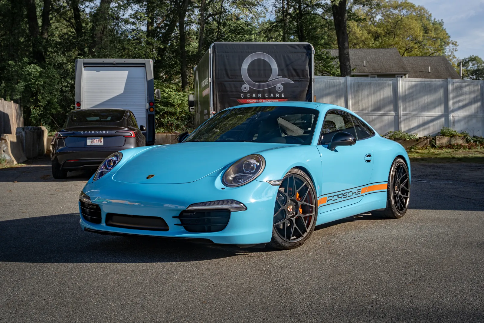 Blue Porsche sports car with black rims parked outdoors near trees and trailers under clear sky.