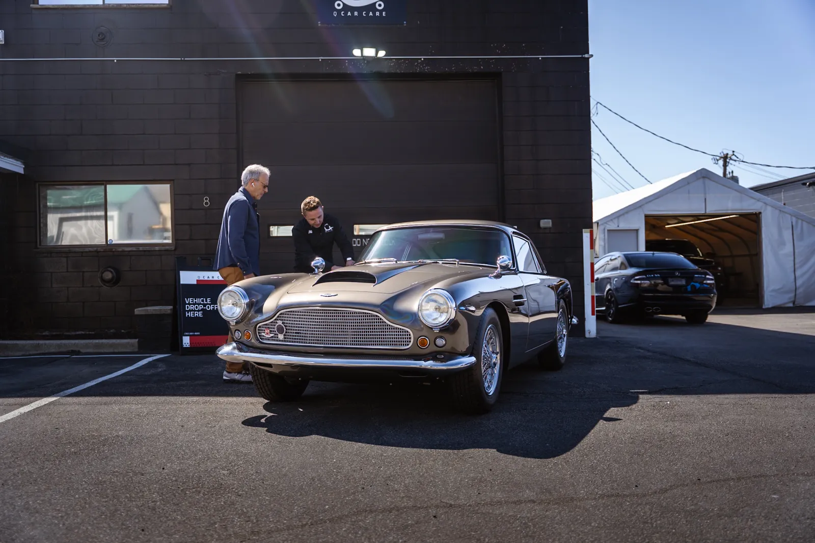 Classic silver Aston Martin parked outside a garage with two men inspecting the vehicle on a sunny day.