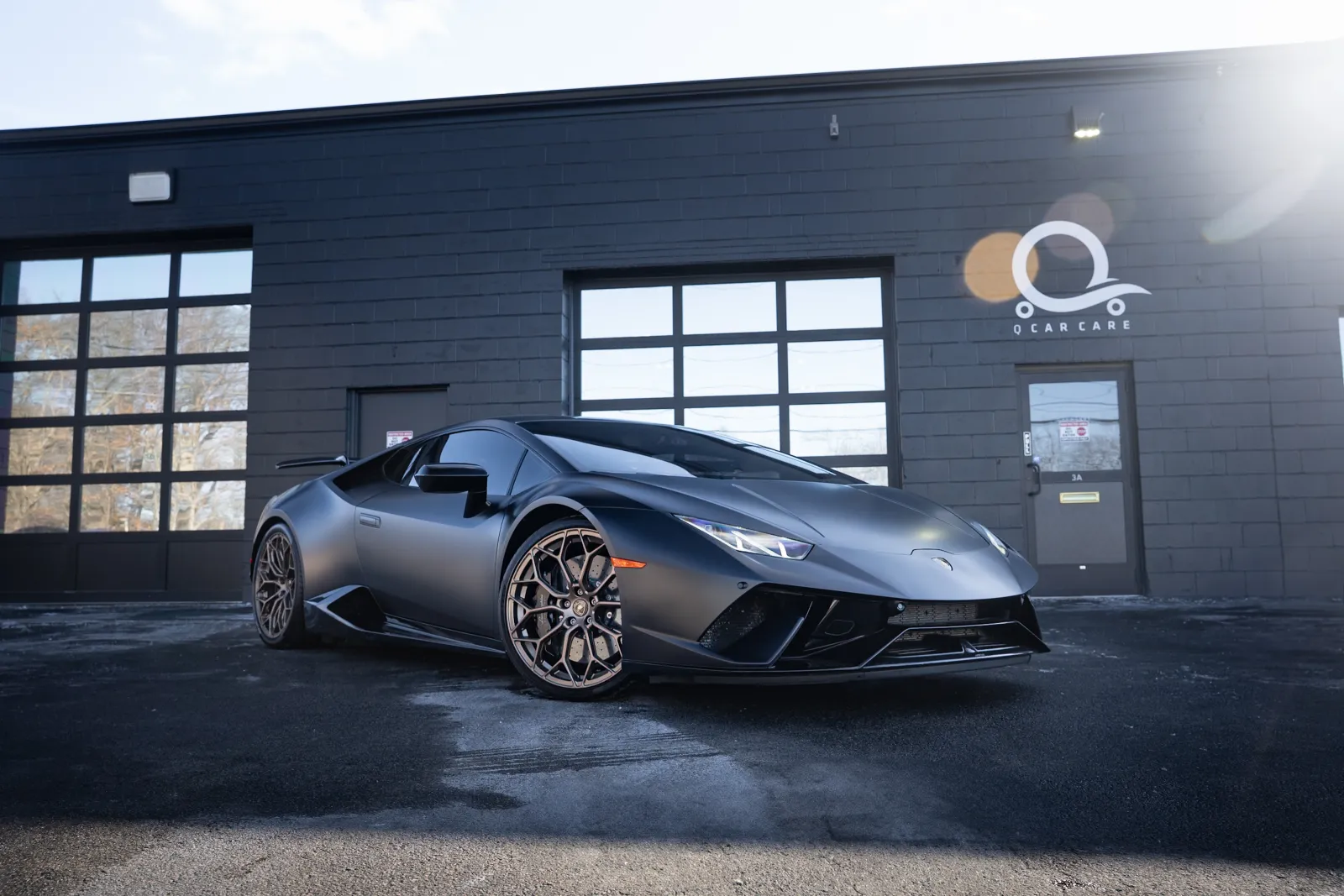 Matte black Lamborghini sports car parked in front of a black garage with glass windows on a sunny day.