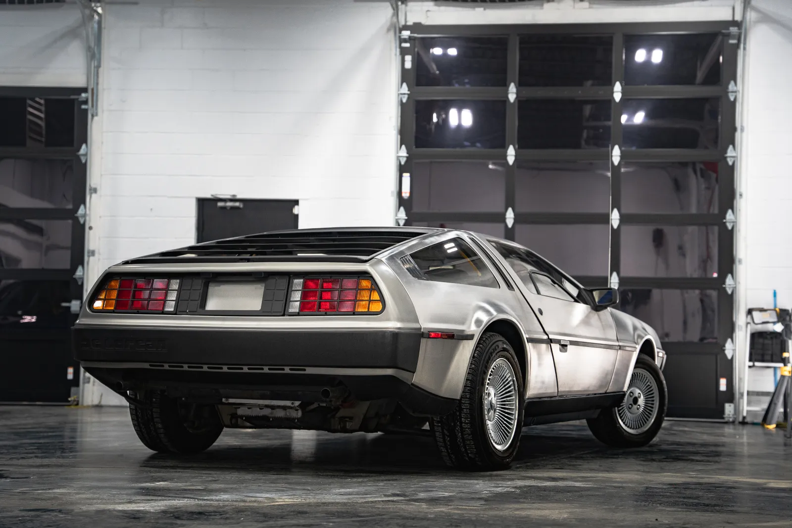 Rear side view of a silver DeLorean DMC-12 vintage sports car in a garage with black doors.