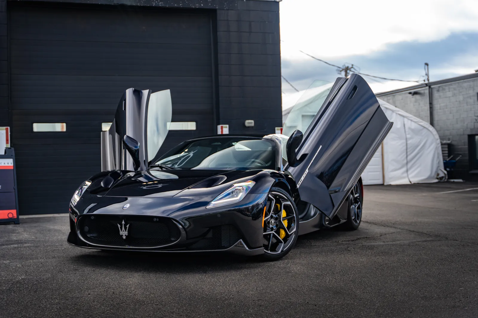 Black Maserati sports car with scissor doors open, yellow brake calipers, parked in front of a garage.