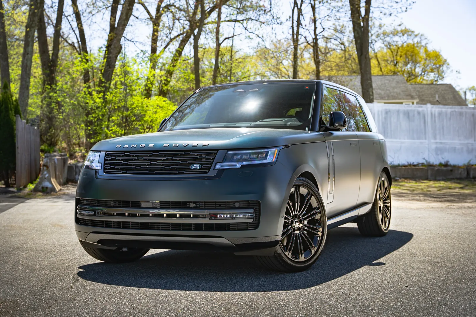 Matte black Range Rover parked on a sunny residential street with trees and fence in the background.