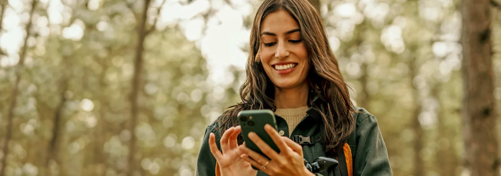 Young woman smiling while using smartphone outdoors in a forest setting during daytime.