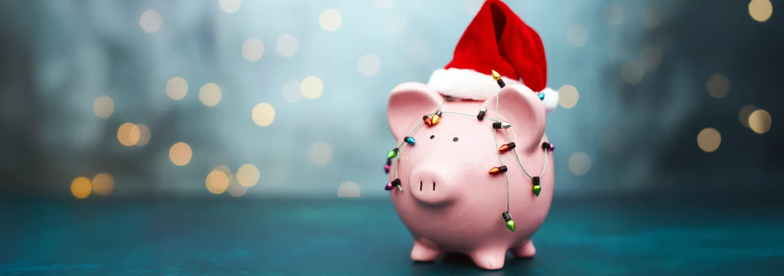 Pink piggy bank wearing a Santa hat wrapped in colorful Christmas lights on blue background with bokeh.