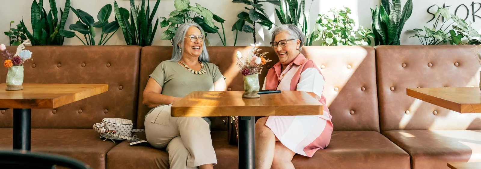 Two elderly women laughing and chatting in a cozy café with plants and warm natural light.