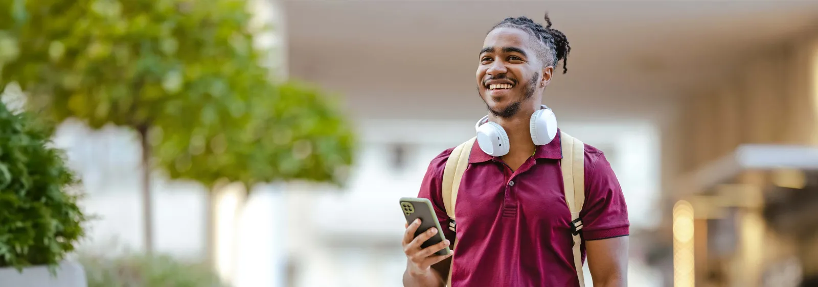 Smiling young man with backpack and headphones holding smartphone outdoors on a sunny day.