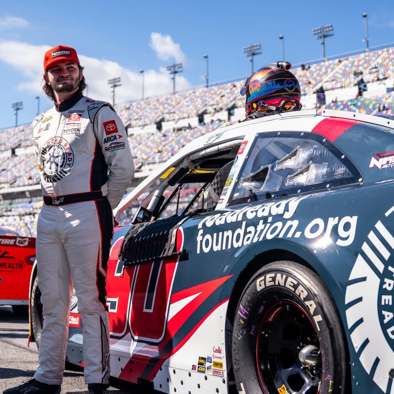 Race car driver in white suit and red cap stands next to black and red number 60 stock car on racetrack