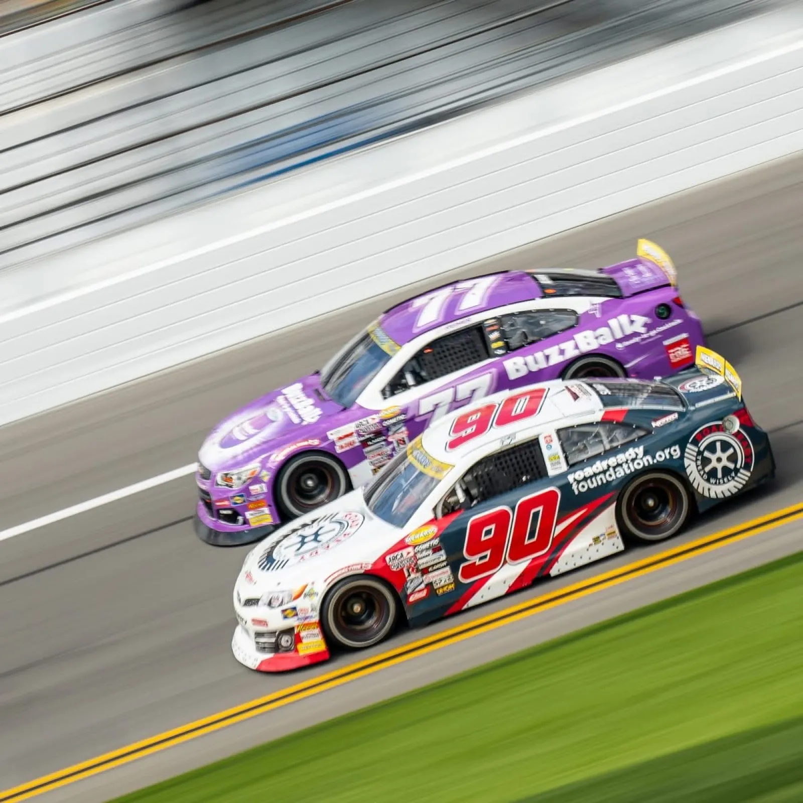 Two stock cars, number 90 in black and white and number 77 in purple, race side by side on a track at high speed.