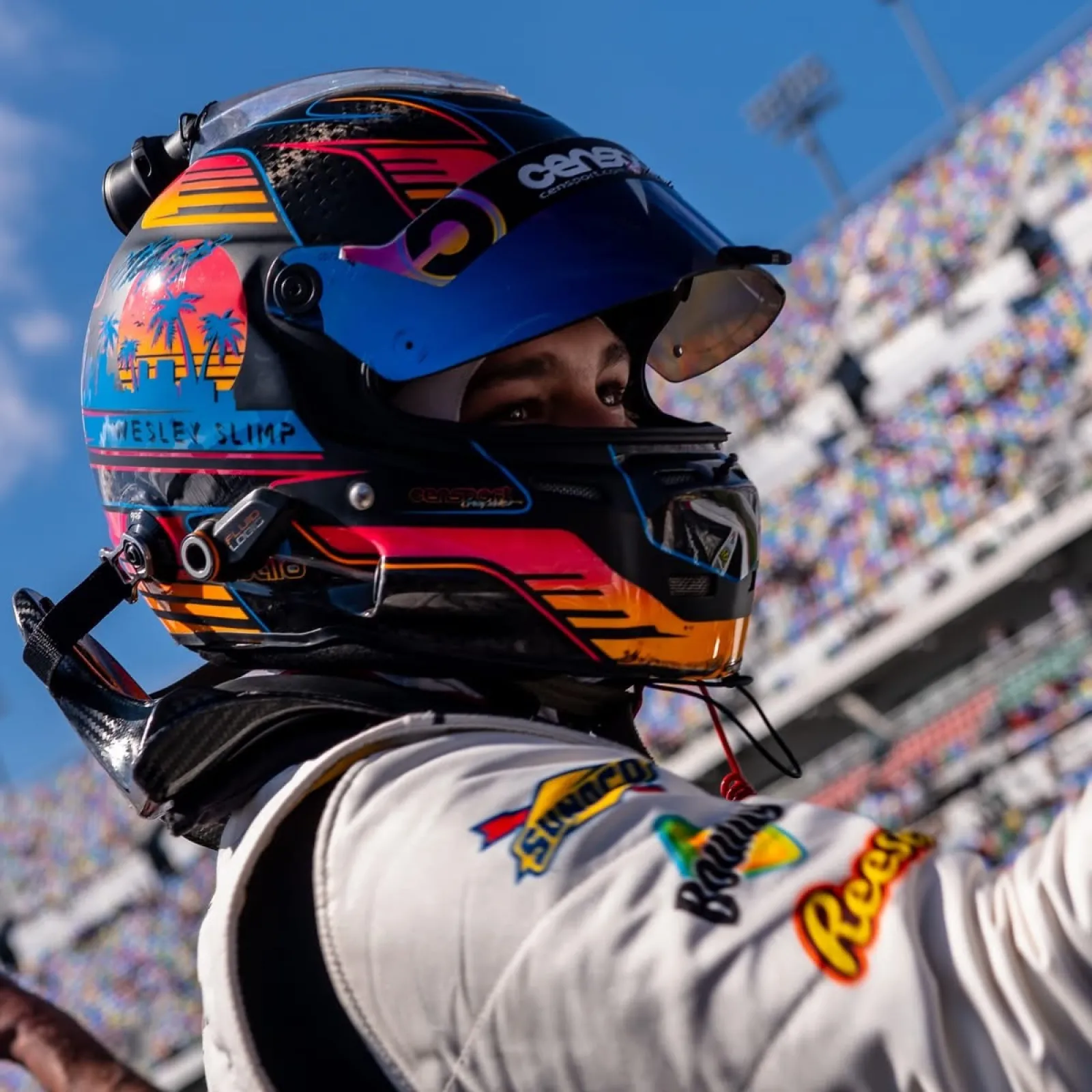 Race car driver wearing a colorful helmet and white suit at a motorsport event with grandstands in the background