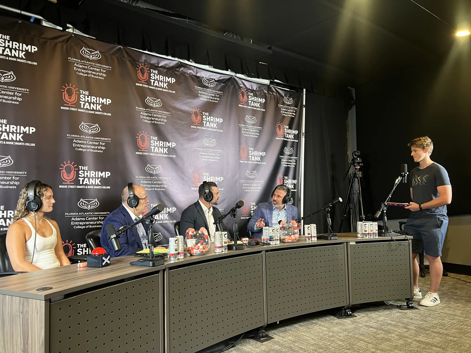 Panel of podcast hosts and guests recording The Shrimp Tank with microphones and a cameraman filming the session.
