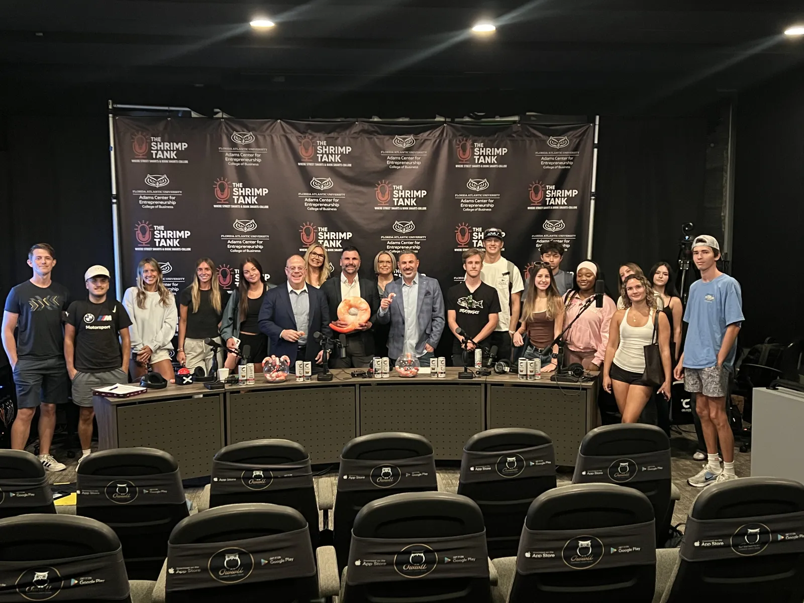 Group of people posing in front of The Shrimp Tank backdrop at entrepreneurship event with a panel table and prizes.