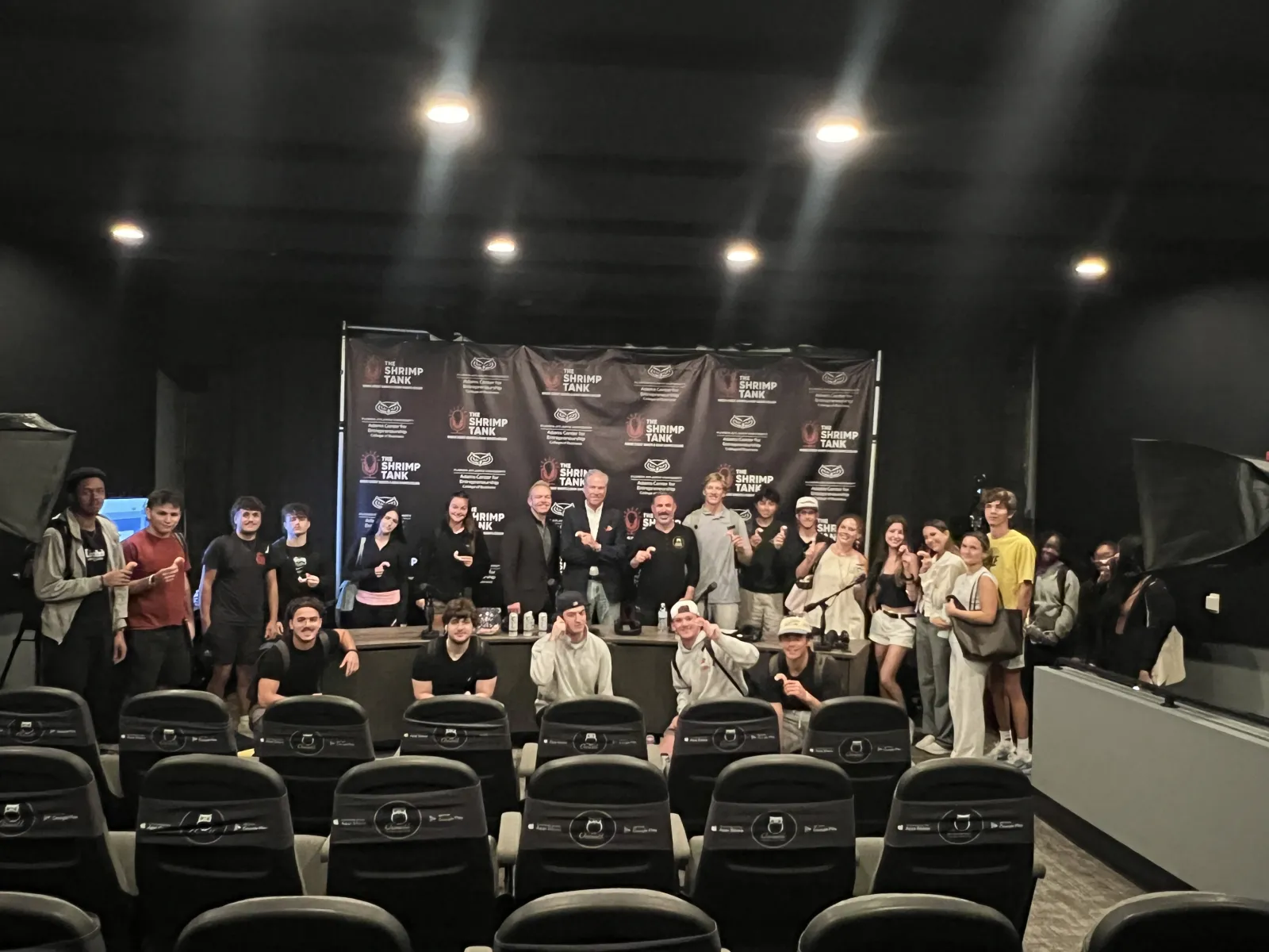 Group of people posing in a theater with Shrimp Tank branded backdrop and empty chairs in the foreground.