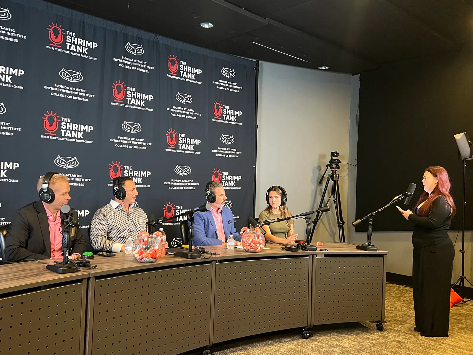 Panel of four people with headphones behind microphones facing a standing woman speaking into a microphone in a recording studio