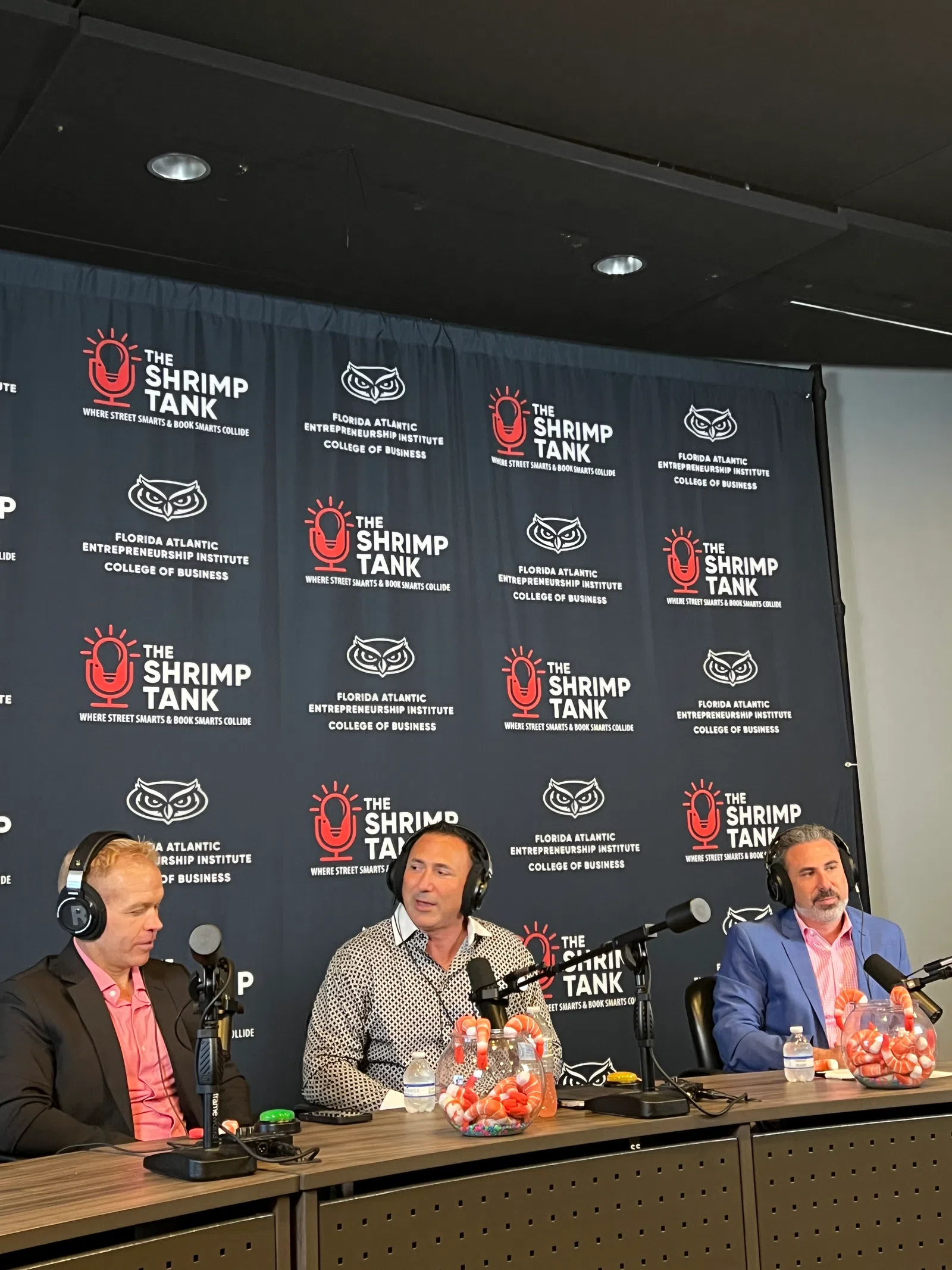 Three men in headphones recording a podcast at a table with The Shrimp Tank and Florida Atlantic backdrop