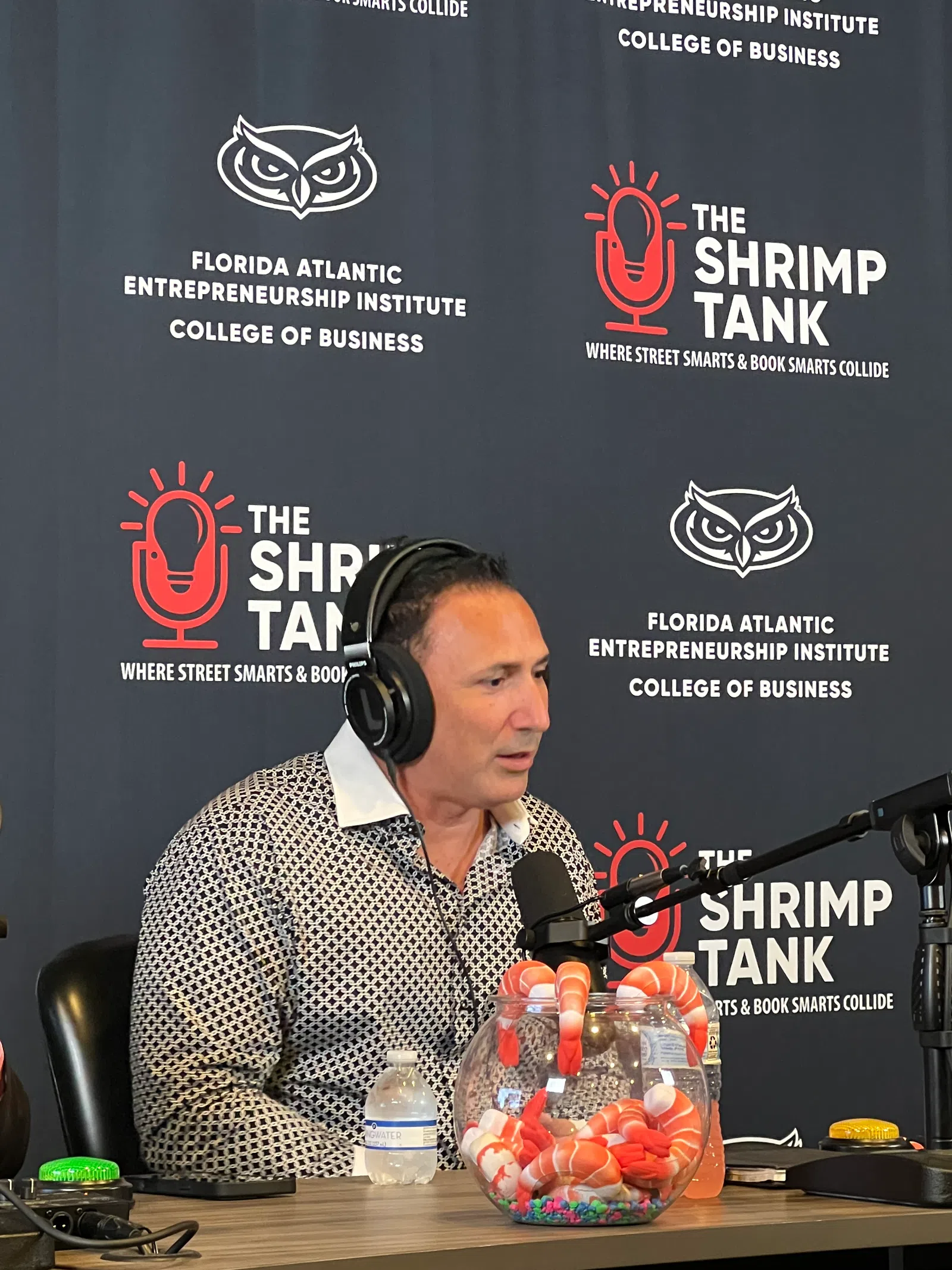 Man wearing headphones speaking into a microphone on The Shrimp Tank podcast with branded backdrop.