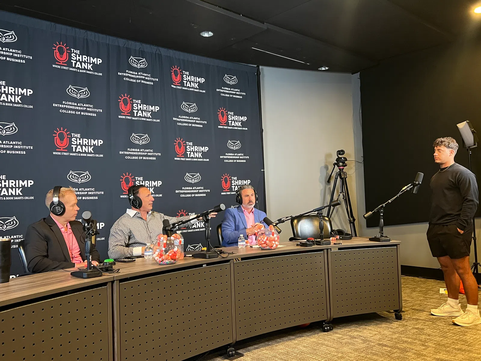Three men seated at a podcast table labeled The Shrimp Tank with a young man standing opposite, recording setup visible.