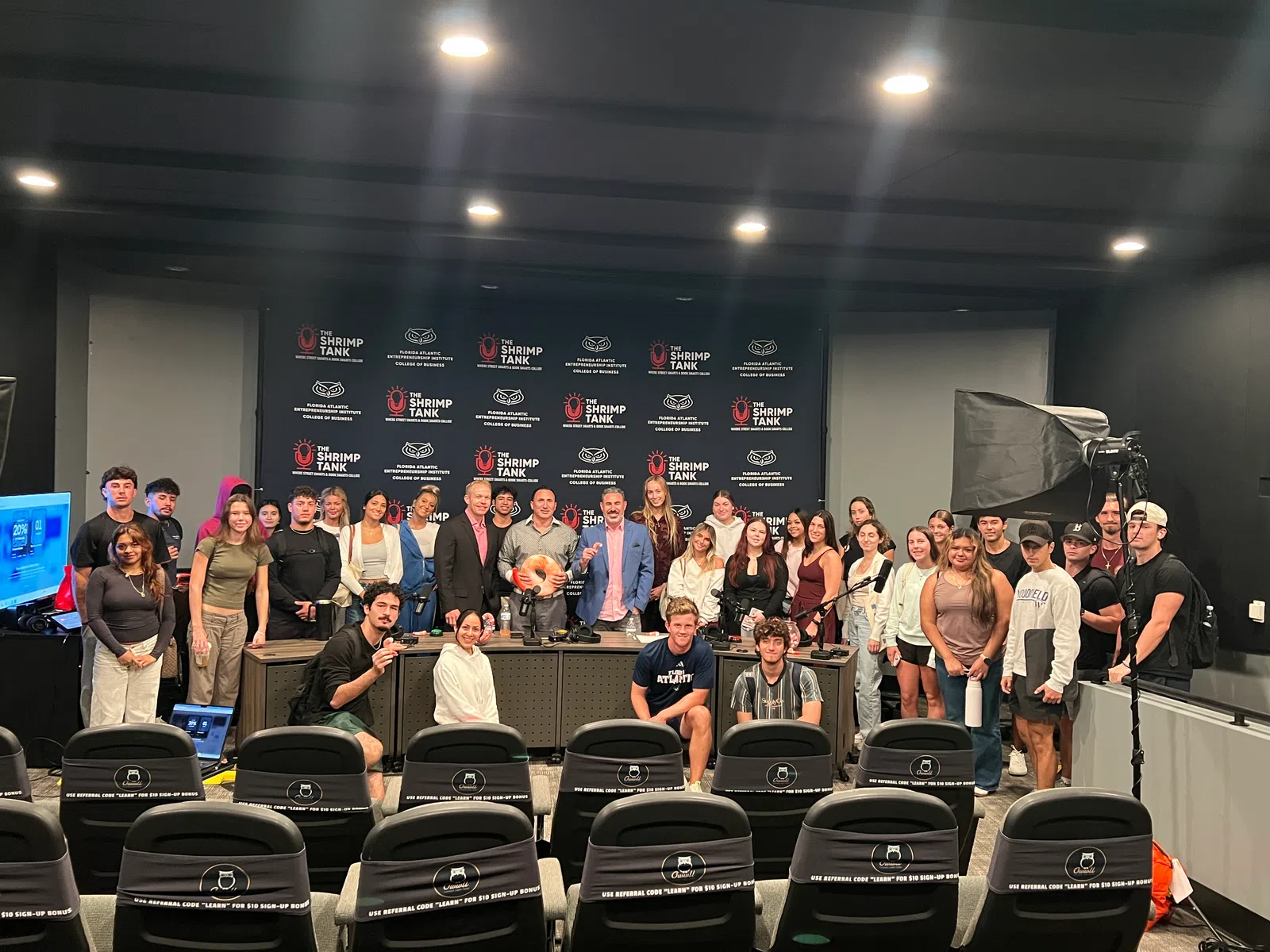 Group photo of diverse individuals in a studio with Shrimp Tank branding and recording equipment.