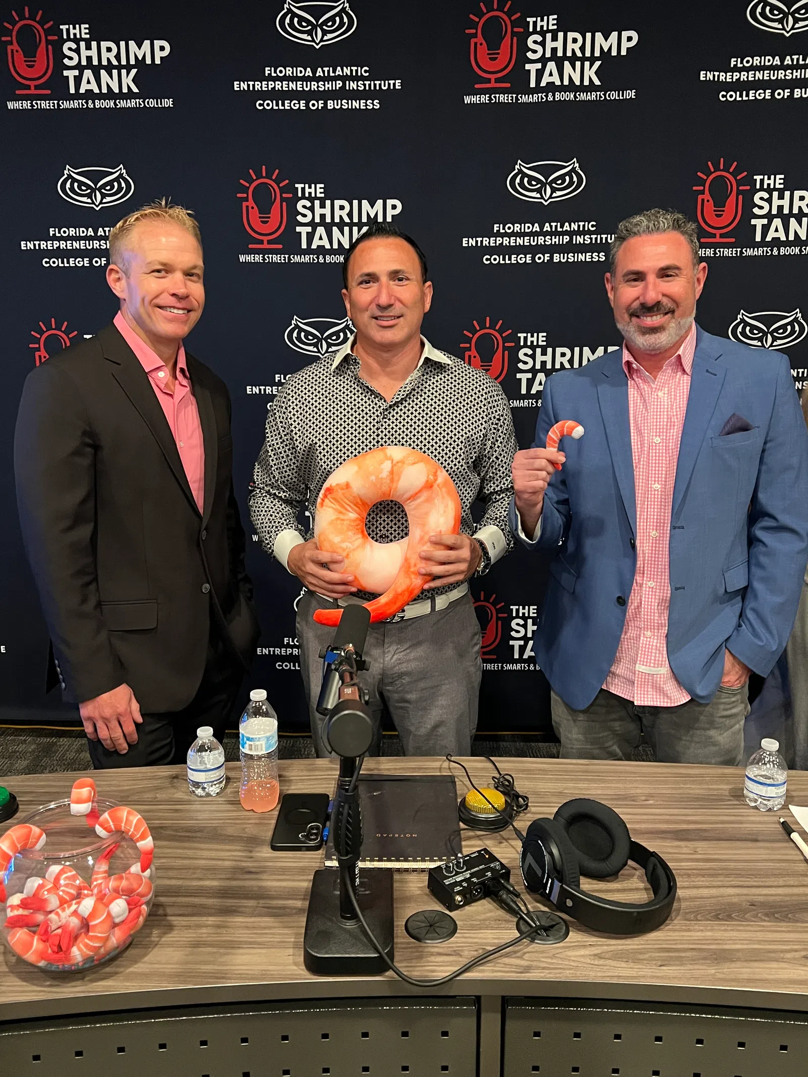 Three men standing behind a table with shrimp props and podcast equipment, with The Shrimp Tank backdrop