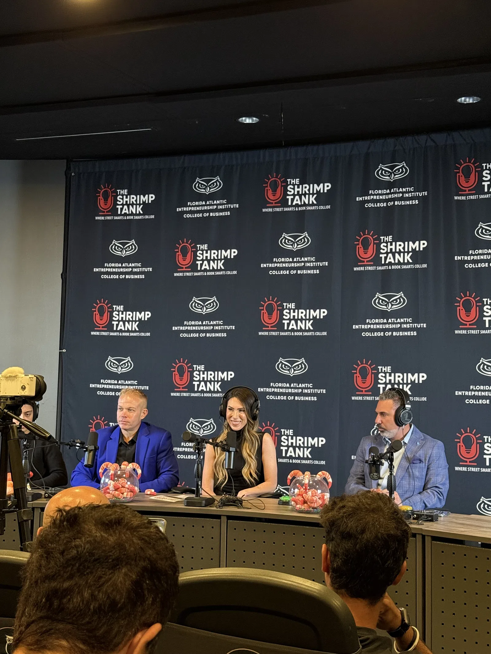 Panel of three hosts with microphones and headphones at The Shrimp Tank event backdrop, engaging audience at conference.