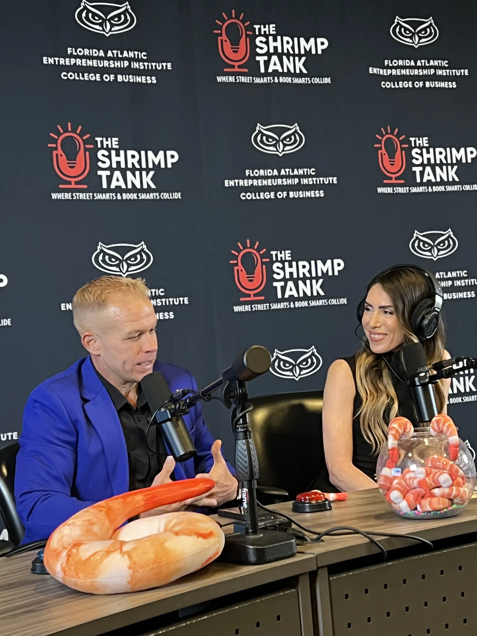 Two hosts recording a podcast at The Shrimp Tank with branded backdrop and shrimp-themed props on the table
