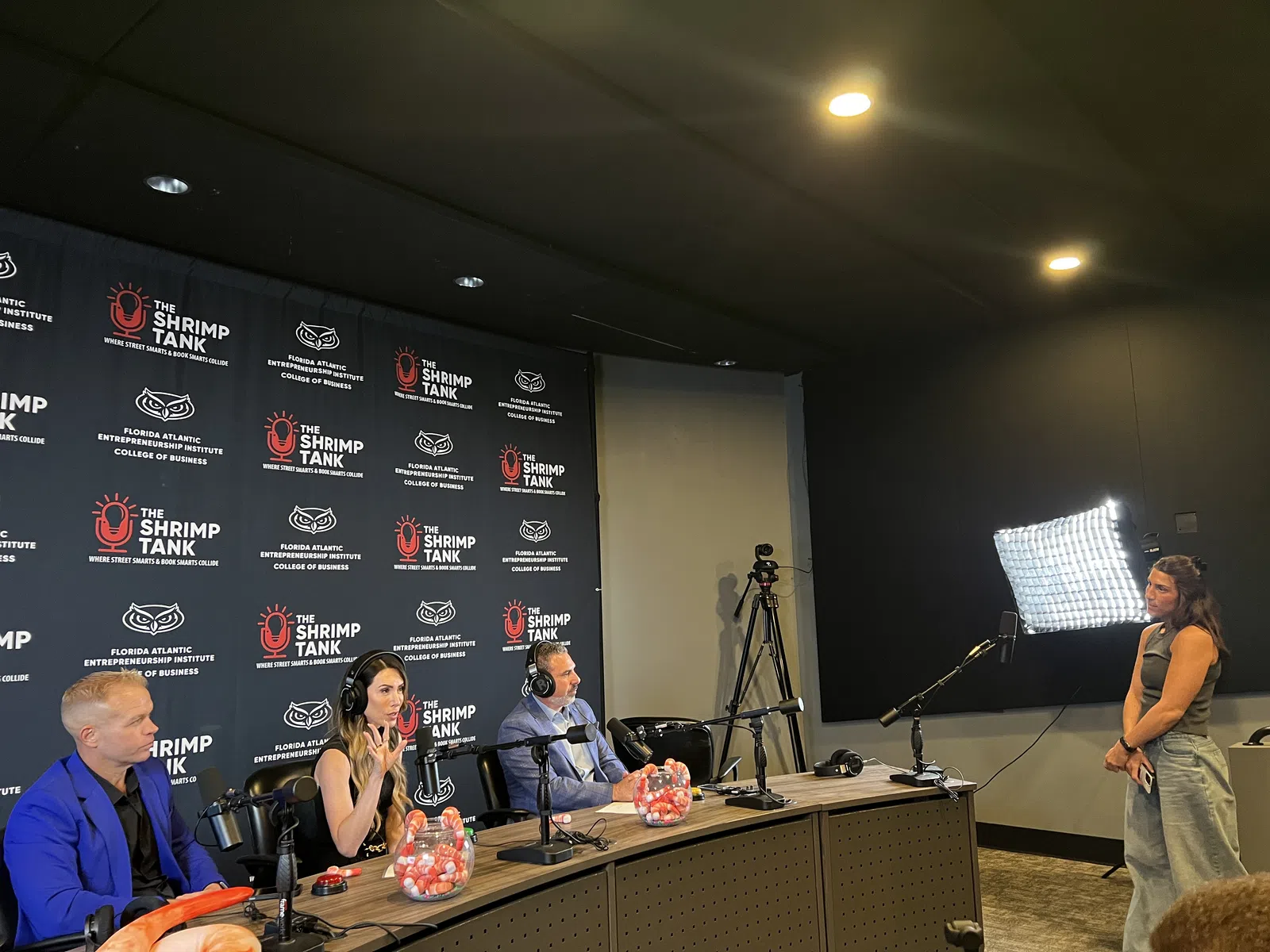 Three judges sit at a panel with microphones and headphones in a podcast studio while a woman stands near a light setup.