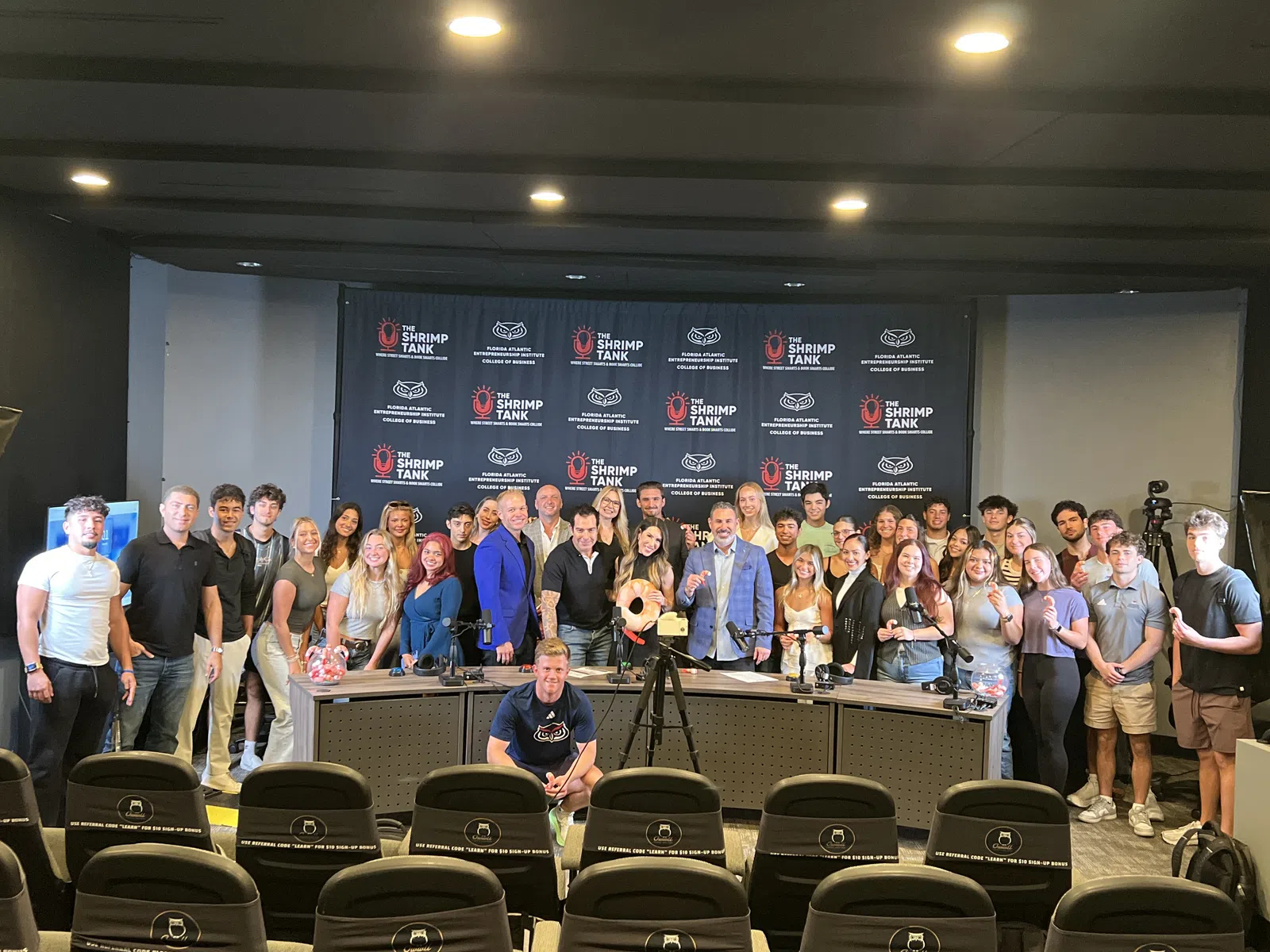 Large group of people posing in front of a backdrop with The Shrimp Tank logo at an event or conference.
