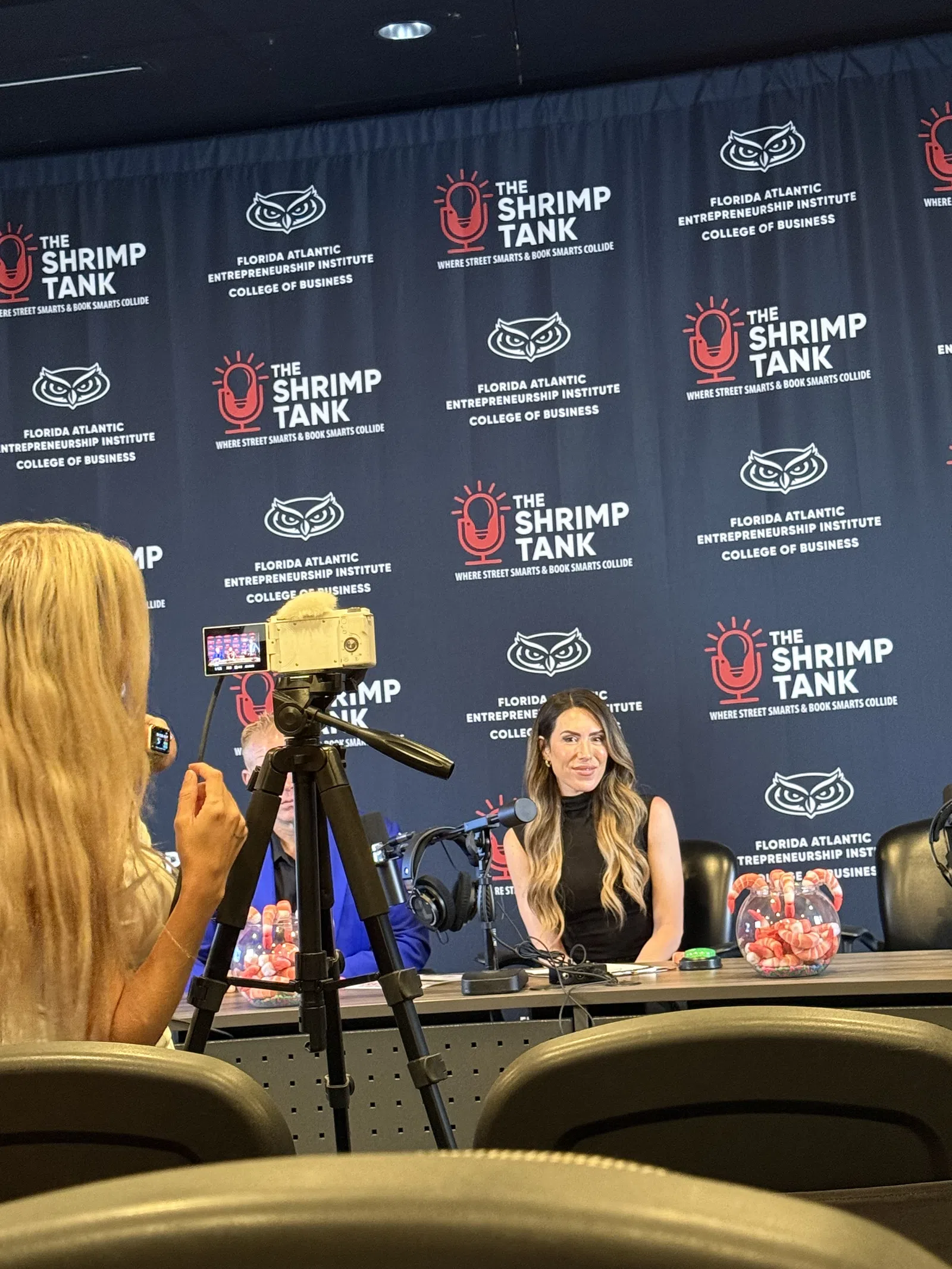 Woman speaking at a Shrimp Tank event with camera and branded backdrop at Florida Atlantic Entrepreneurship Institute.