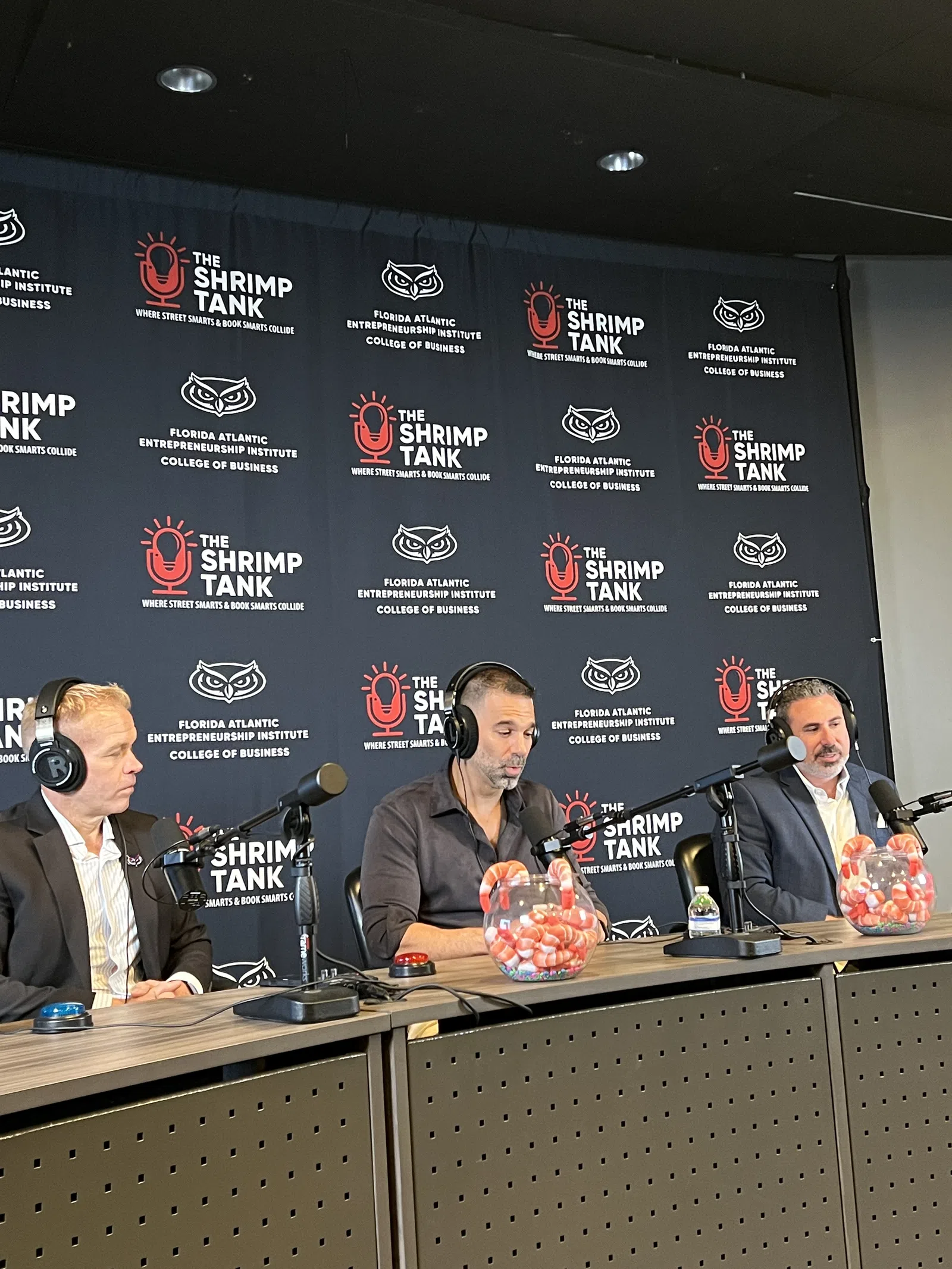Three men wearing headphones speaking into microphones at The Shrimp Tank event backdrop at Florida Atlantic University.