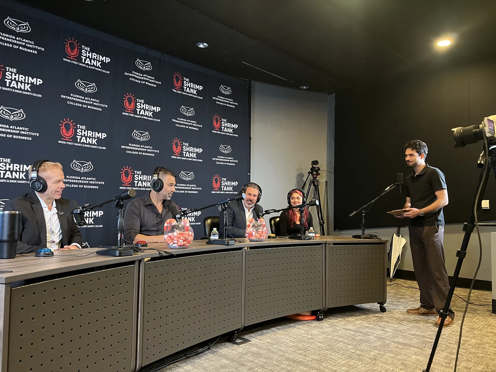 Panel of four judges in a podcast studio discussing on microphones with a host standing near a camera setup.