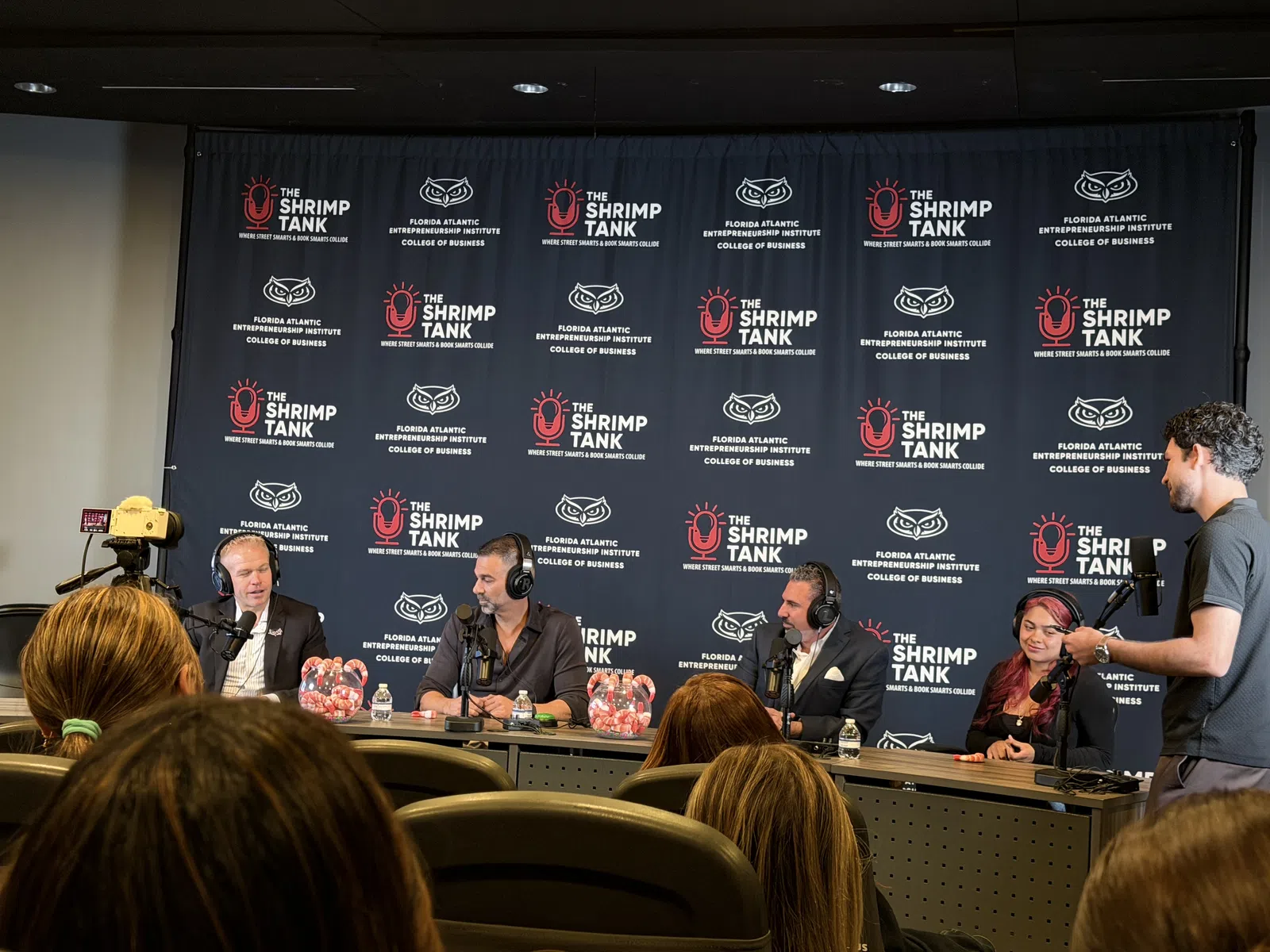 Panel of speakers at The Shrimp Tank event with branded backdrop and audience in foreground
