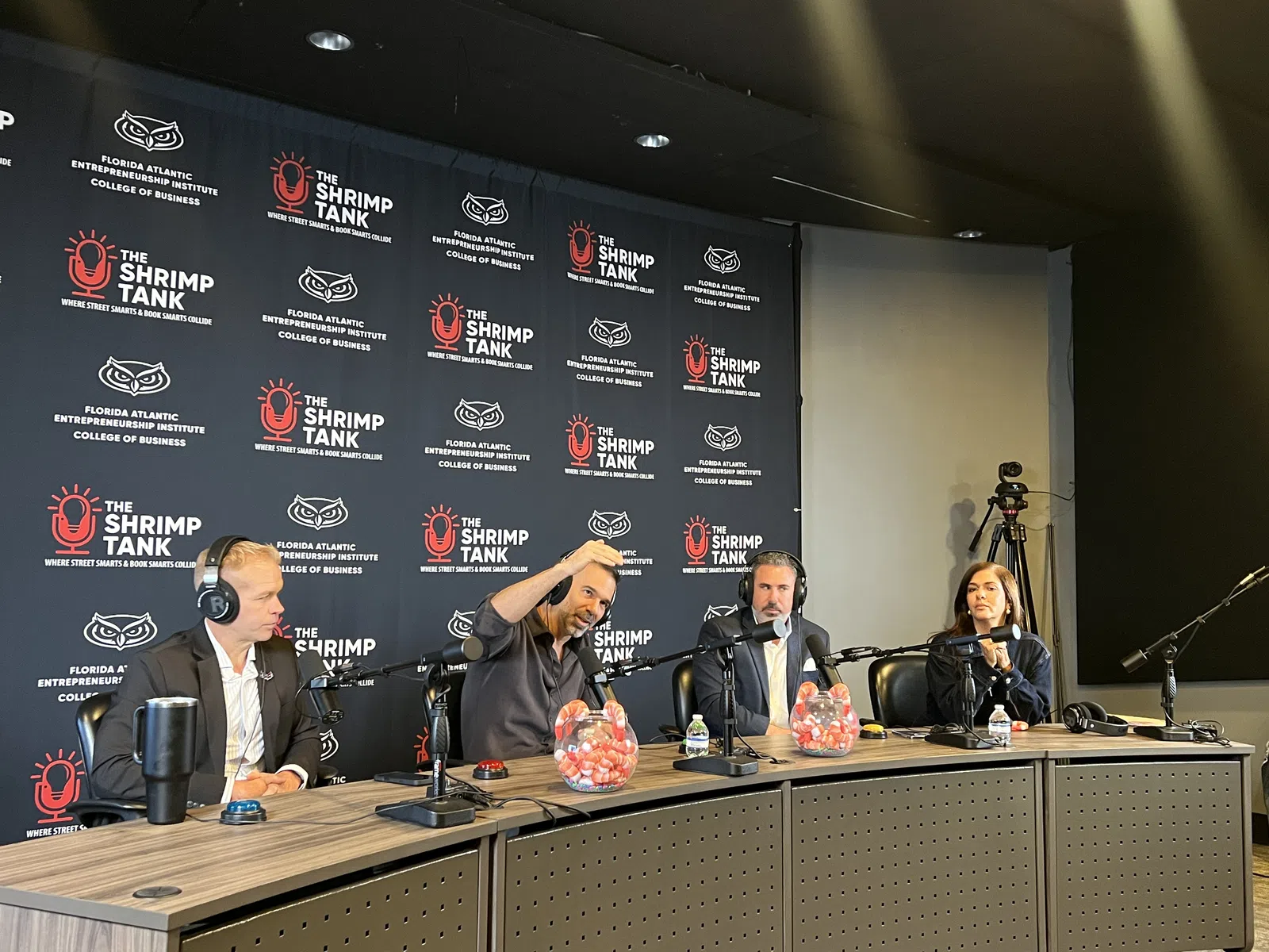 Panelists wearing headphones participate in a podcast recording at The Shrimp Tank event with branded backdrop.