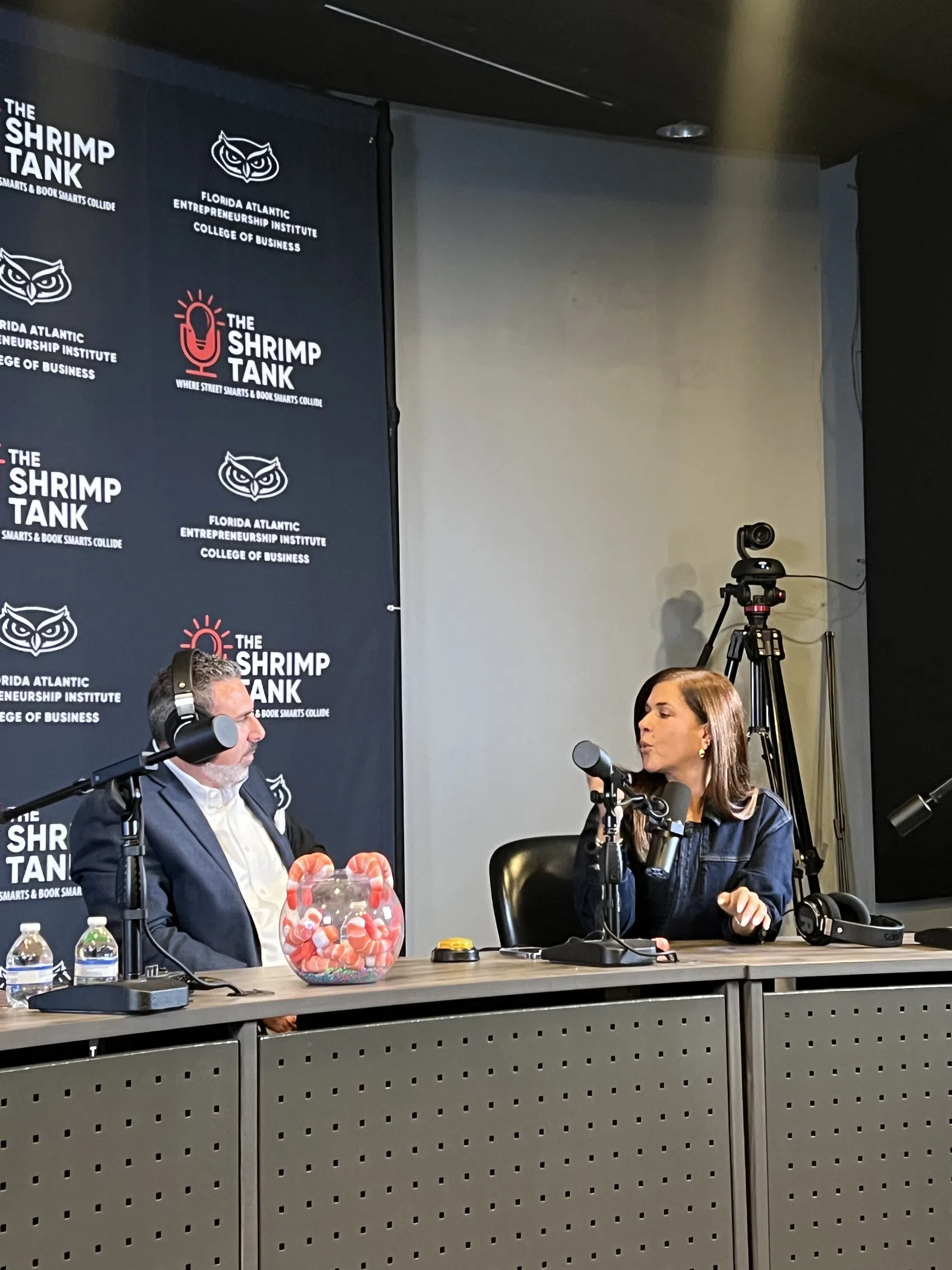Two people recording a podcast episode at a table with microphones and a backdrop for The Shrimp Tank show.