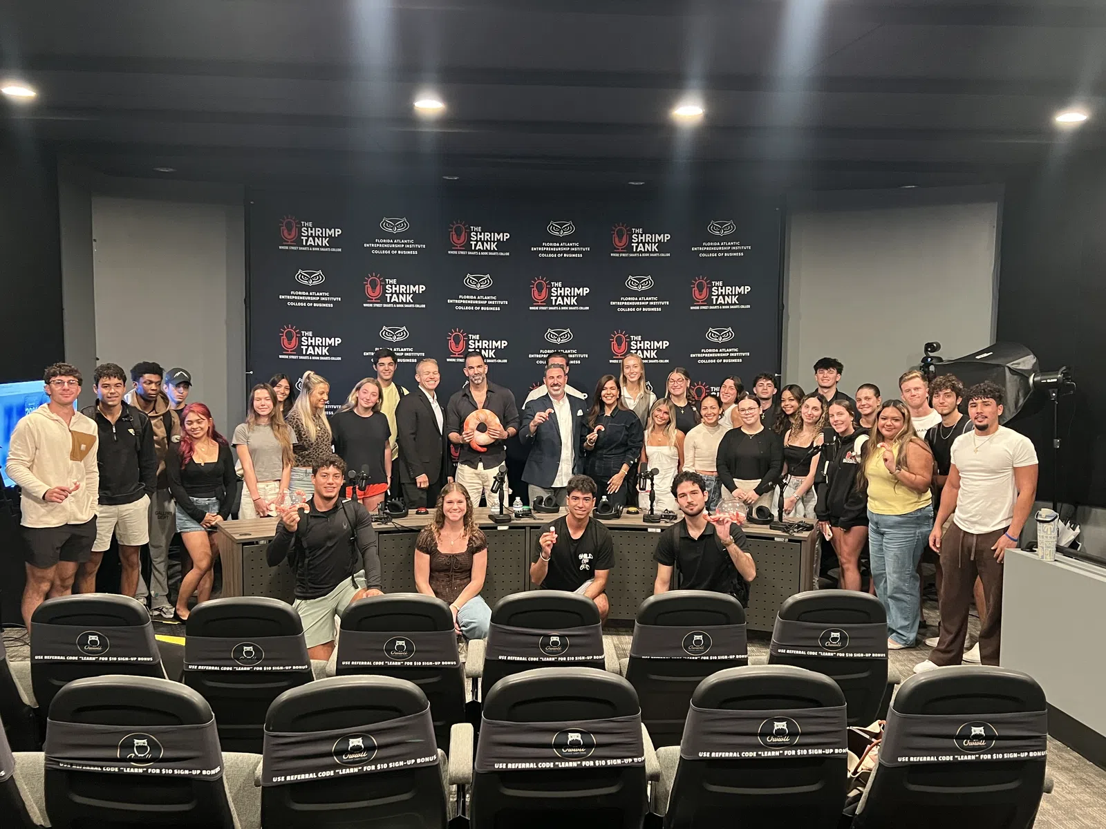 Group photo of diverse young adults and presenters at The Shrimp Tank event in a studio with branded backdrop and seating.